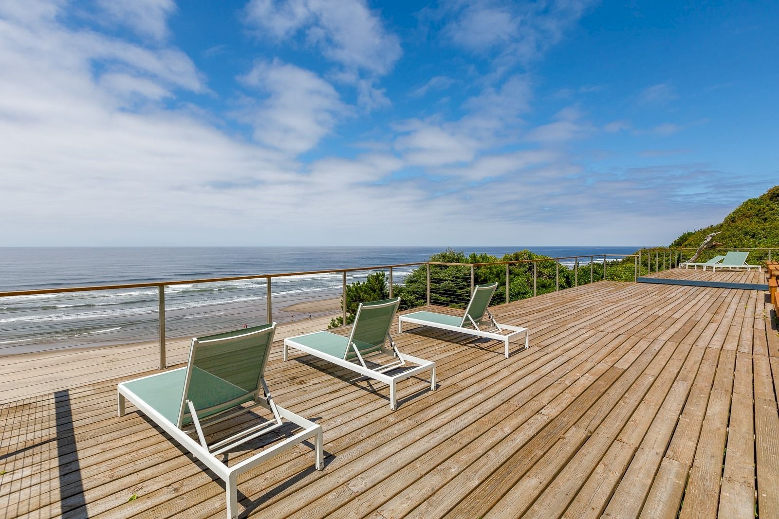 A wooden oceanfront deck with railings and four green lounge chairs under a bright blue sky with clouds.