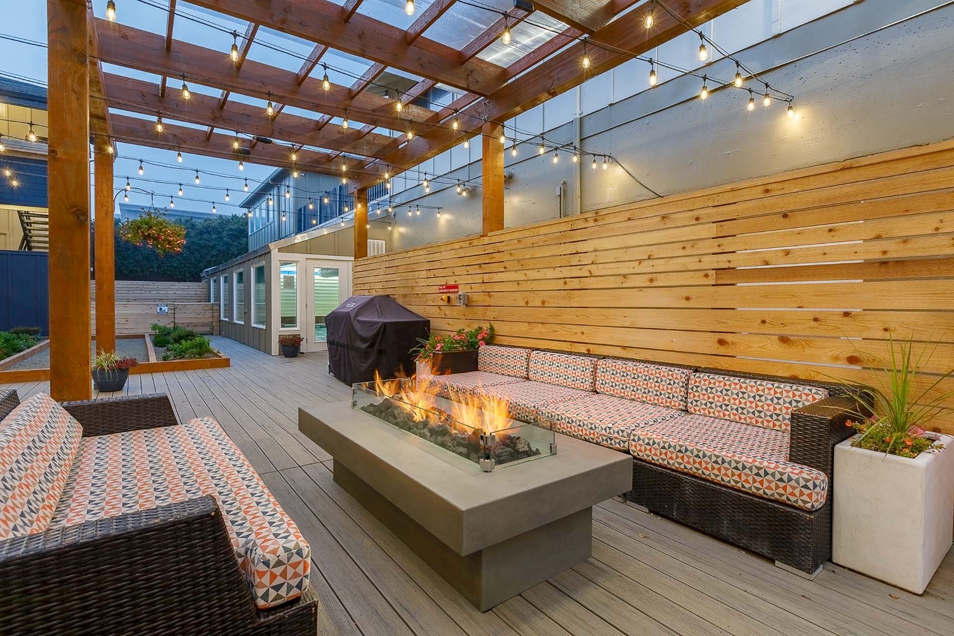 A cozy outdoor patio with wooden pergola, string lights, cushioned seating, and a gas fire pit for warmth and ambiance.