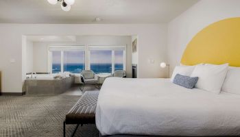 A modern hotel bedroom with a large bed, lounge chairs, bright windows with ocean view, and a yellow accent wall.