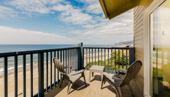 A beachfront balcony with lounge chairs and a small table, looking out over sandy waves and a cloudy blue sky.