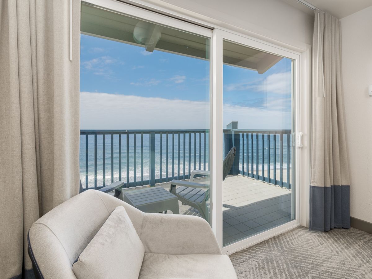 A bright seaside room with a sofa and large sliding doors opening to an ocean view balcony with railings and patio chairs.