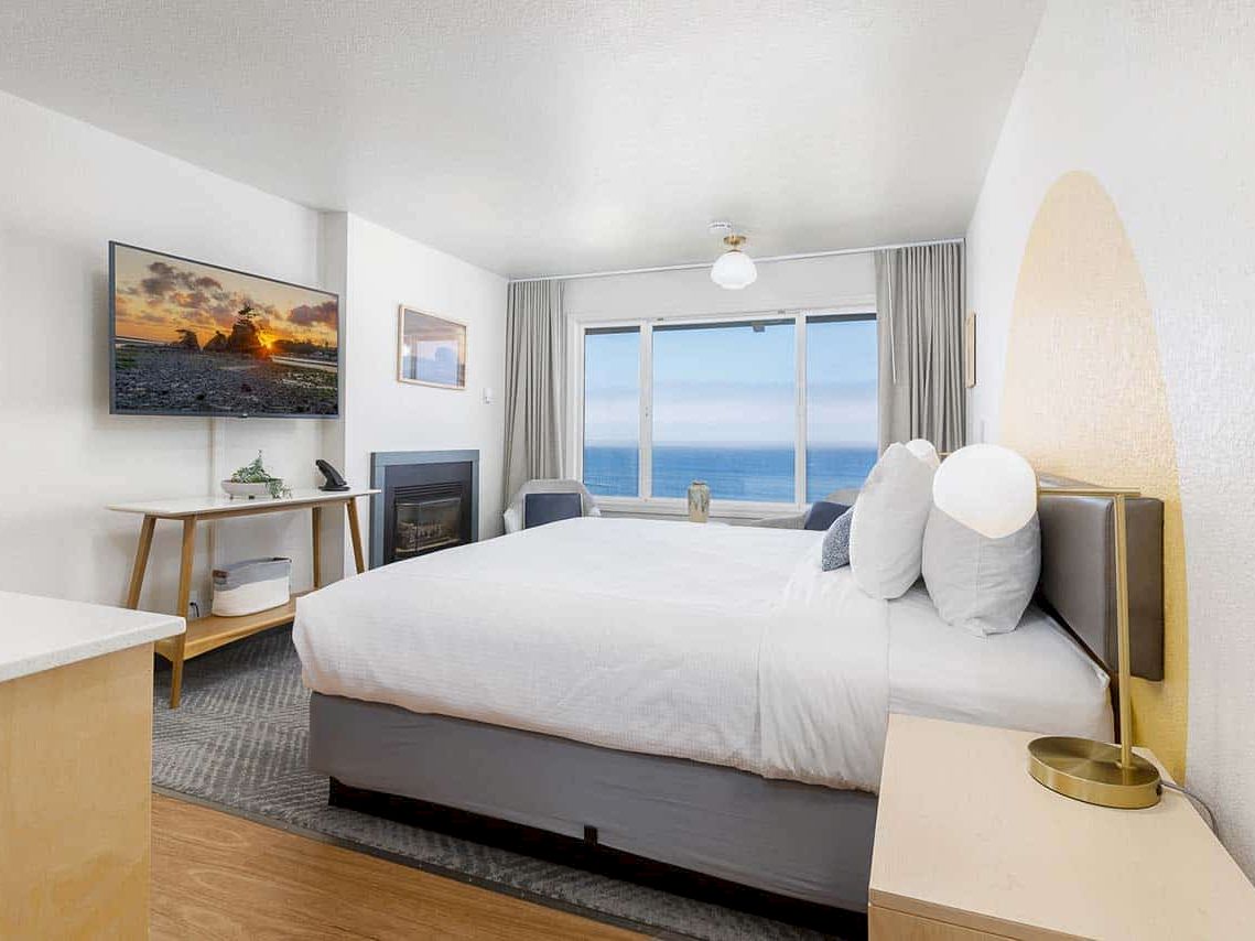 A modern hotel bedroom with a large bed, fireplace, and big window showing an ocean view at sunset.