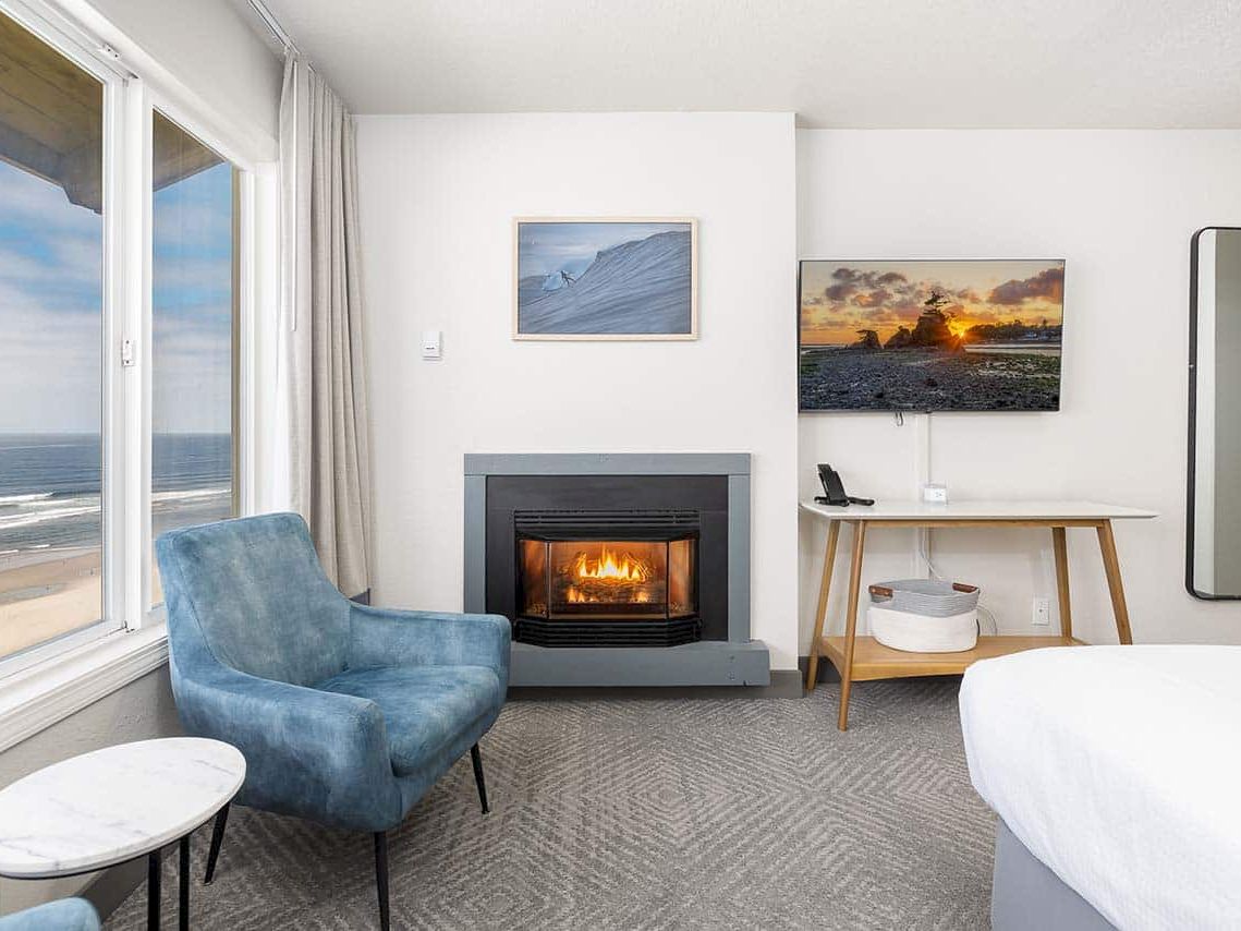 A bright seaside hotel room with a lit fireplace, blue chair, desk, ocean view window, and framed artwork.