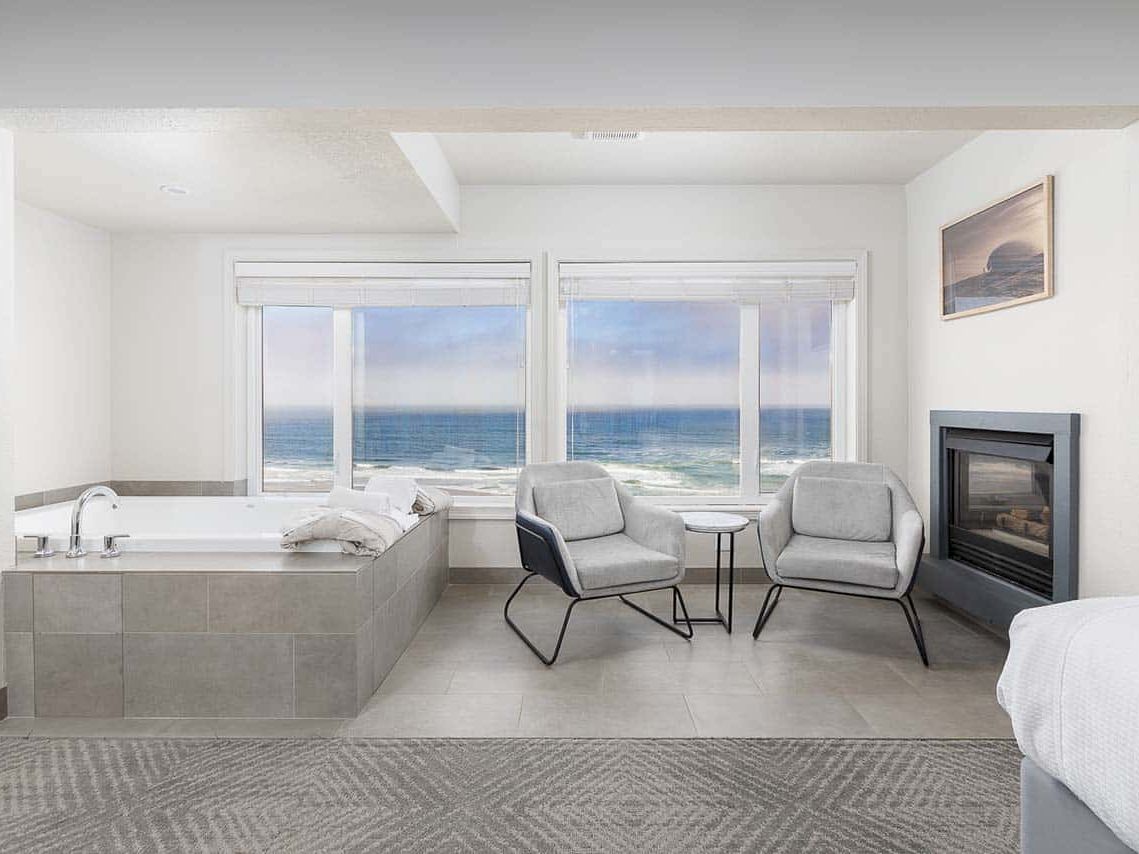 Bright modern room with ocean view, two chairs, small table, fireplace, and a built-in bathtub.