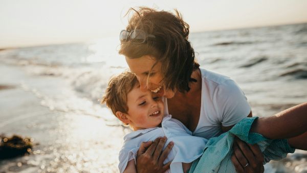 A woman in sunglasses hugs a smiling child on a sunny beach, with waves and sparkling sea in the background.