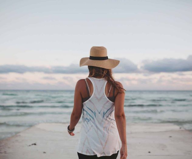 A woman in a straw hat and white dress stands on a beach by the ocean at dusk.