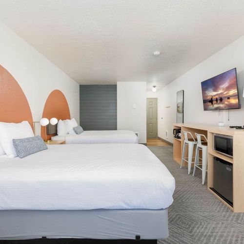A modern hotel room with two beds, a desk and TV, a lamp, and neutral decor in view, ending in a cozy layout.