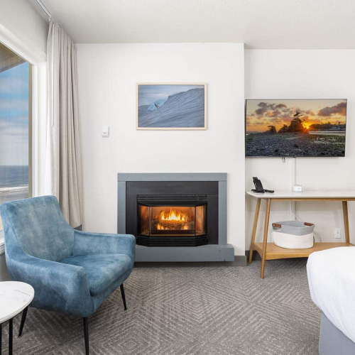 A cozy hotel room with a fireplace, beach-view window, framed wall art, a desk with TV, and a lounge chair.