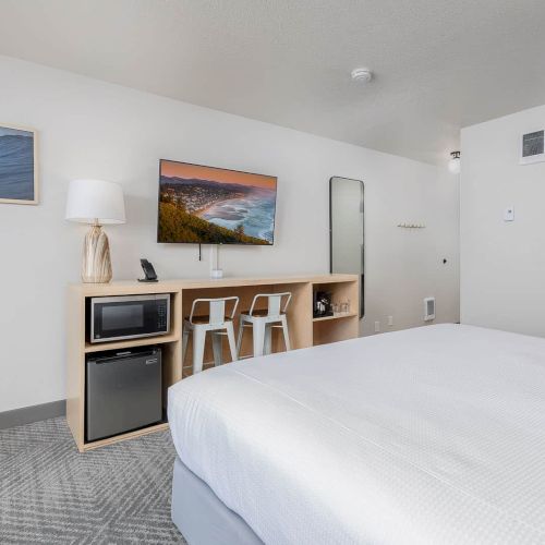 A modern hotel room with a large bed, desk with microwave and fridge, wall art, and an open doorway.