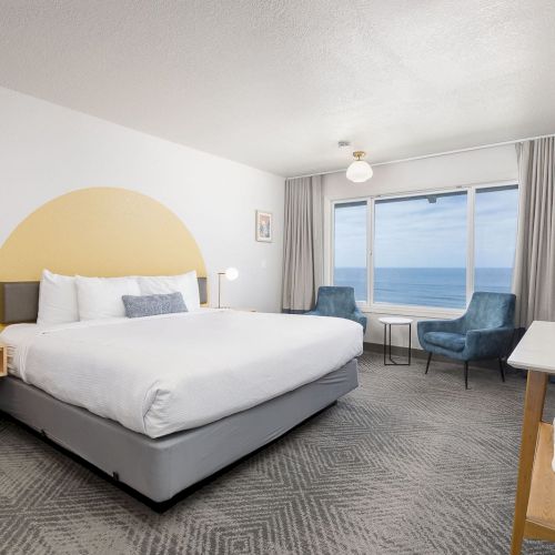 A modern hotel bedroom with a large bed, blue chairs, ocean-view window, and wall-mounted TV.