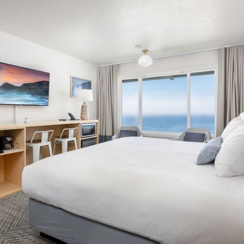 A bright hotel room with a large bed, TV, minibar cabinet, and curtains framing a view of the ocean.
