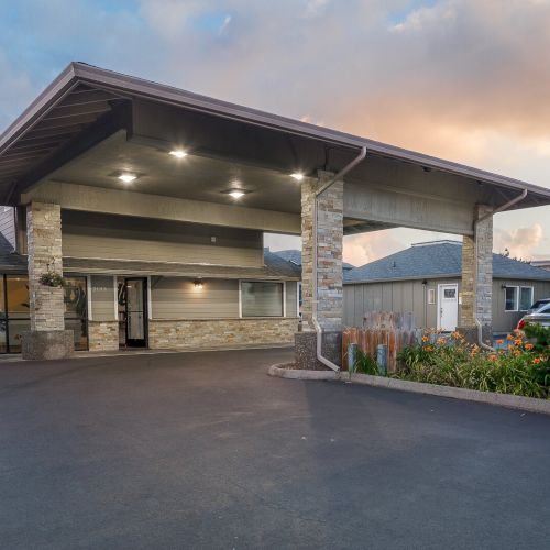 A modern building entrance with a covered carport, stone pillars, landscaped flowers, and parked cars under an evening sky.