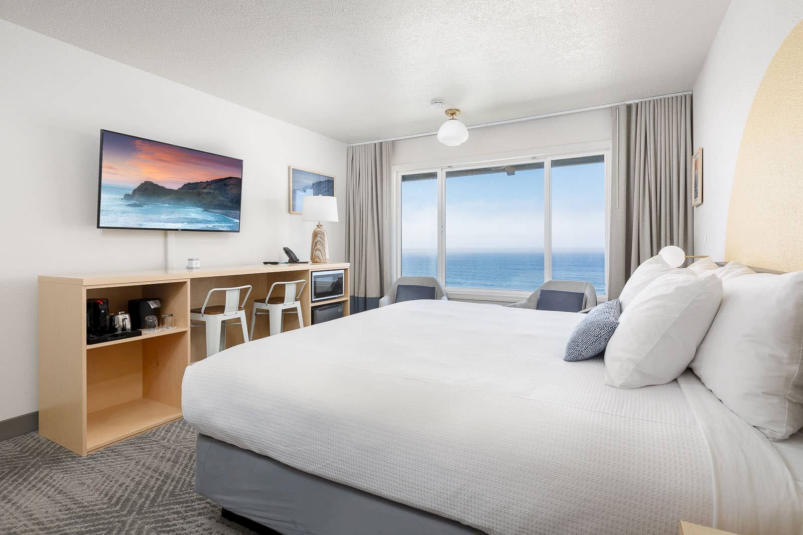 Bright hotel-style bedroom with a white bed, desk coffee setup, wall TV, and large windows showing the ocean view.