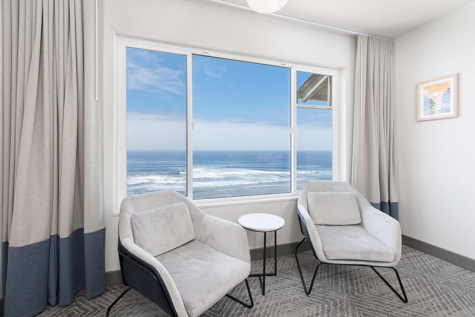 Bright seaside room with two armchairs and a small table by a large window showing waves outside.