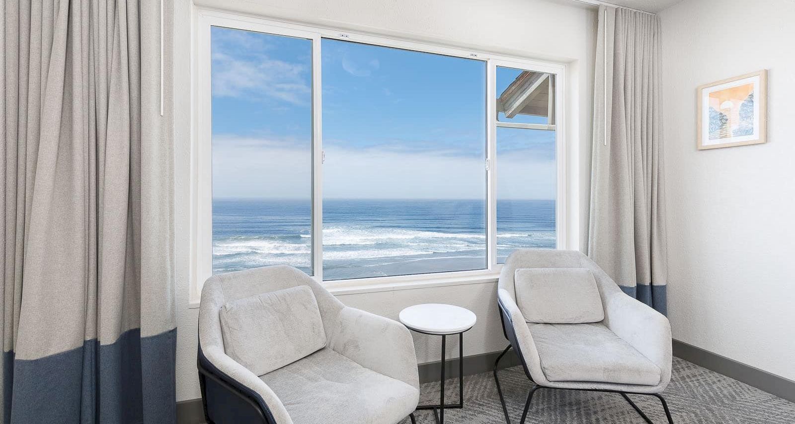 Bright seaside room with two armchairs and a small table by a large window showing waves outside.