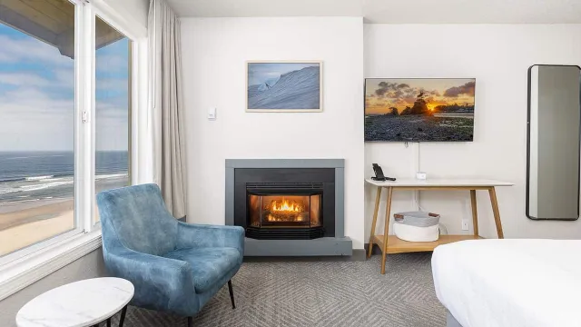 A modern seaside hotel room with ocean-view window, lit fireplace, blue armchair, desk, artwork, TV, and a bed.