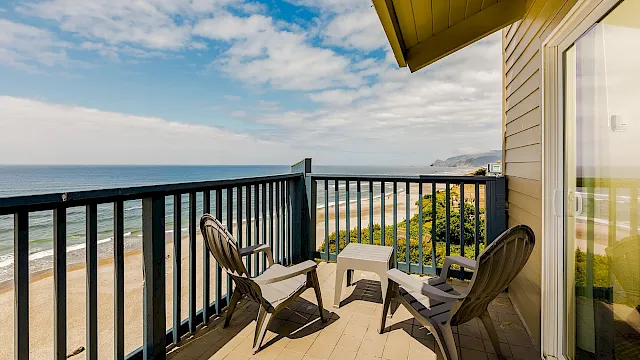 Balcony with patio chairs and umbrella, overlooking sandy beach and ocean under a partly cloudy blue sky.